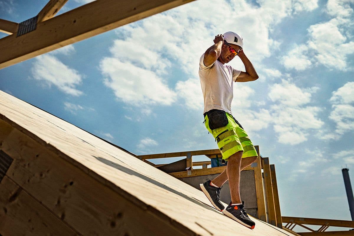 Mannlig håndverker går nedover et tak i ført caps, solbriller, hvit t-skjorte og gul arbeidsshorts