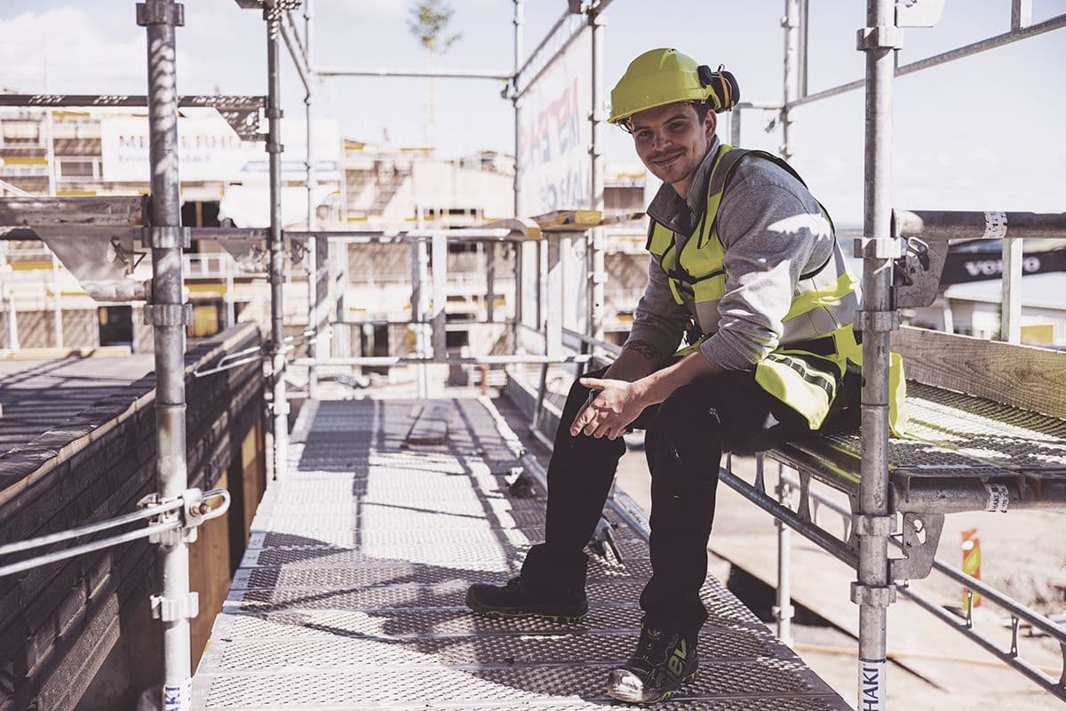 smilende håndverker sitter i et stillas og ser inn i kamera