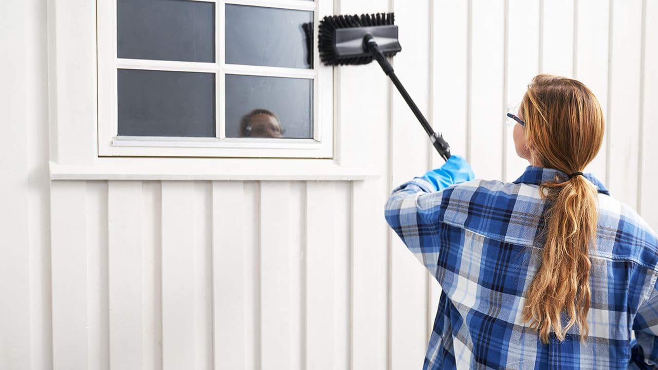 Kvinne som skrubber fasaden på huset med skurekost før maling utvendig.