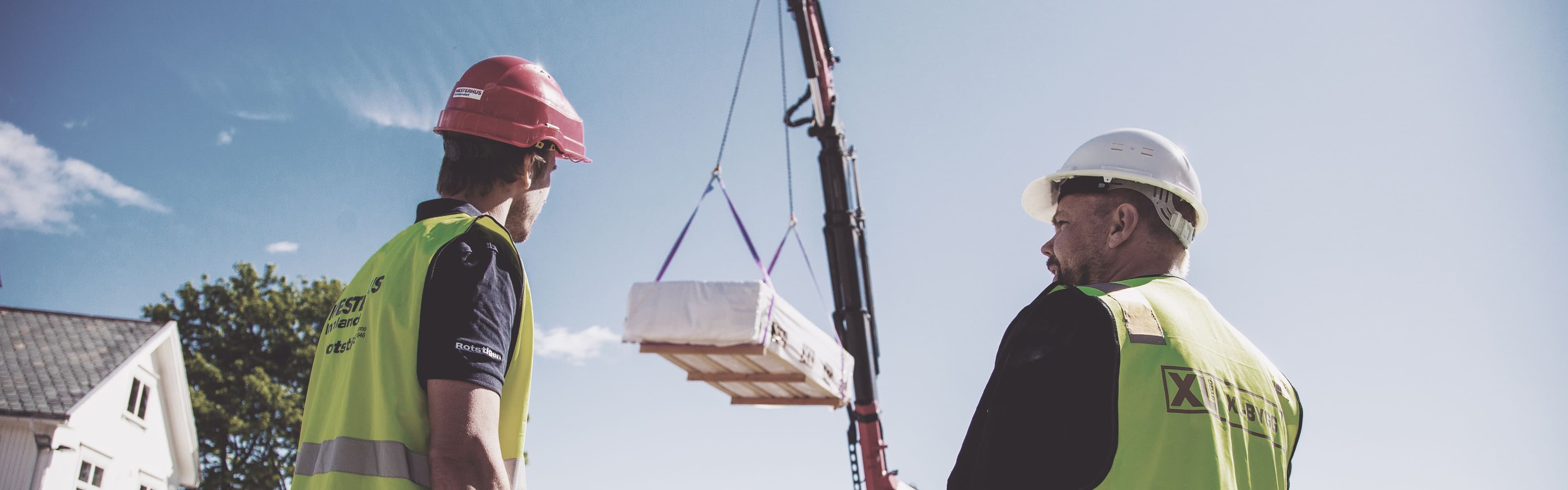 to menn med hjelm står på en byggeplass og ser på materialer som heises opp