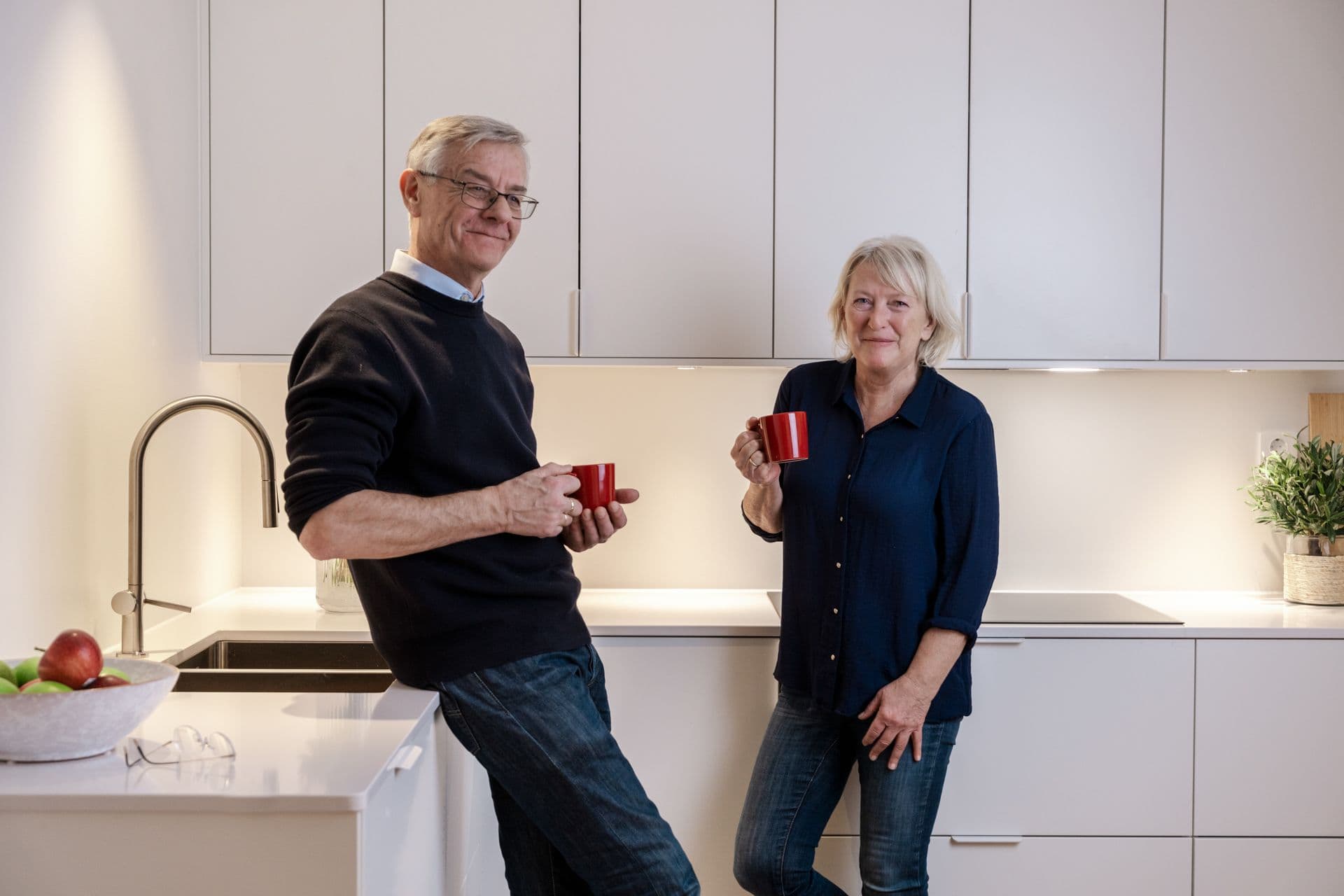 Mann og kvinne som står i et moderne hvitt kjøkken med røde kaffekopper.
