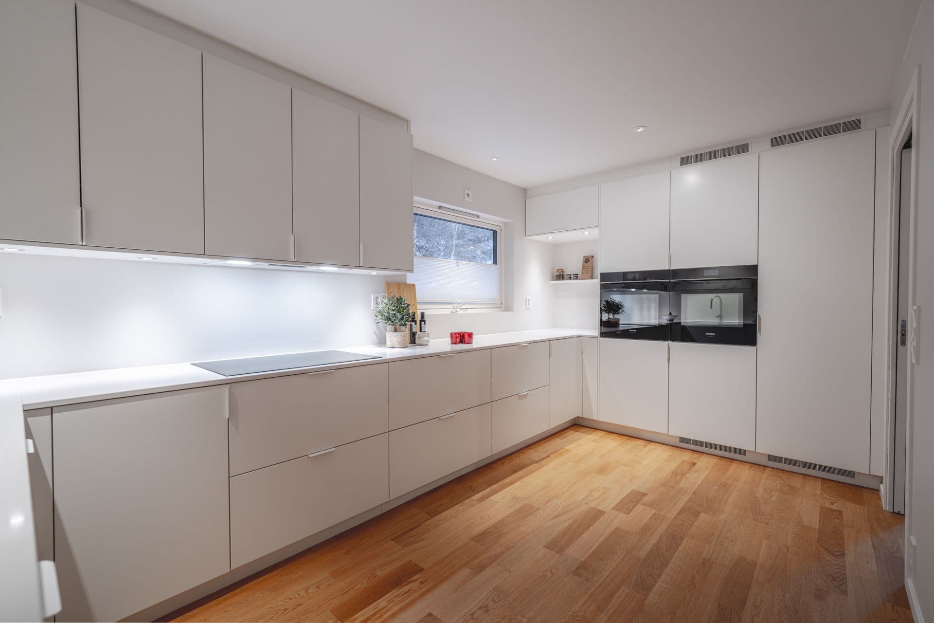 Moderne hvitt kjøkken med hvit benkeplate med høyskapsvegg og eikeparkett.